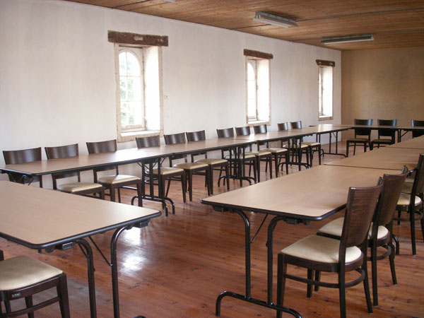 Photo d'une salle de réunion de capacité de 50 personnes avec des tables tout autour du centre de la salle formant un carré et des chaises autour de ce grand carré de tables par terre rouge plafond platre blanc et fenetres sur un grand coté puis un tableau pour écrire et les projections