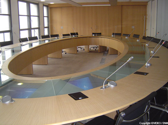 Photo d'une salle de réunion de capacité de 50 personnes avec des tables tout autour du centre de la salle formant un carré et des chaises autour de ce grand carré de tables par terre rouge plafond platre blanc et fenetres sur un grand coté puis un tableau pour écrire et les projections