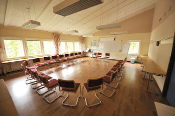 Photo d'une salle de réunion de capacité de 50 personnes avec des tables tout autour du centre de la salle formant un carré et des chaises autour de ce grand carré de tables par terre rouge plafond platre blanc et fenetres sur un grand coté puis un tableau pour écrire et les projections